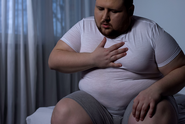 Overweight man sat on bed holding his chest - Sleep Apnoea and Obesity | Sleeptest.co.uk